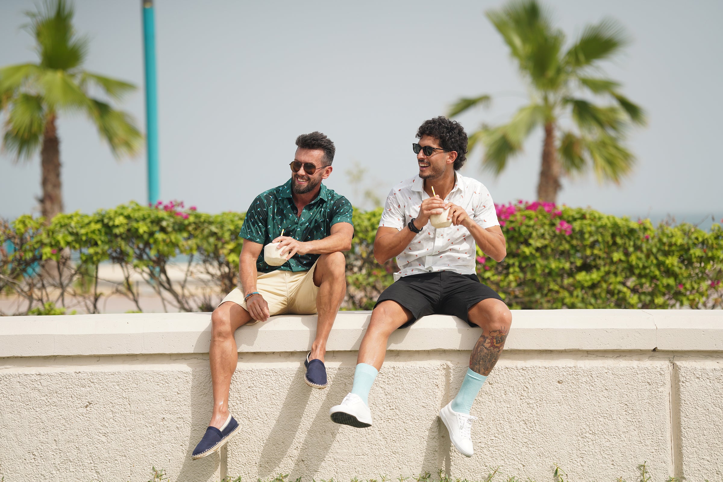 two men in casual attire sitting on a wall, wearing shorts and shirts, engaged in conversation. happy bay